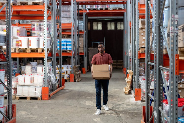 Young male worker of warehouse carrying boxes person walking through warehouse carrying a cardboard box